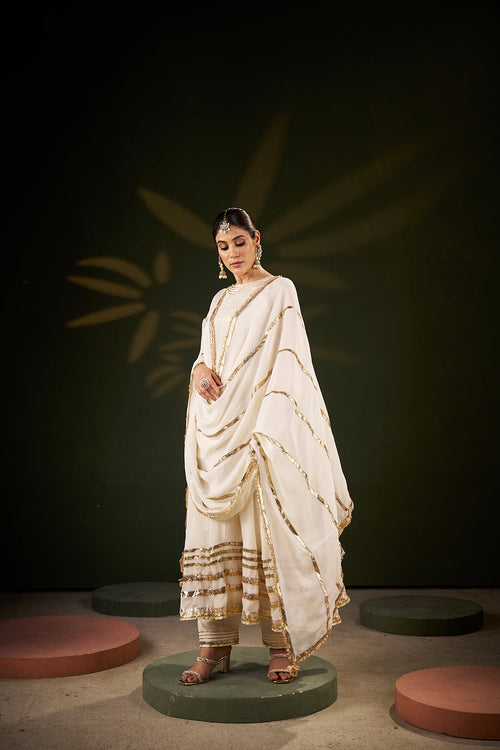 women in White Anarkali and chiffon dupatta with gotta laces standing in a dark background 